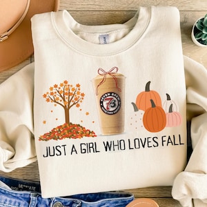 Può includere: Felpa color crema a tema autunnale. Il design include un albero con foglie arancioni, zucche e una tazza di caffè. Il testo sulla maglietta recita "JUST A GIRL WHO LOVES FALL."