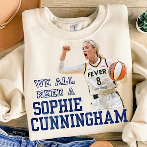May include: Cream-colored sweatshirt with the text "WE ALL NEED A SOPHIE CUNNINGHAM" in navy blue. The sweatshirt features a basketball player in a Fever jersey holding a basketball. The jersey number is 8.
