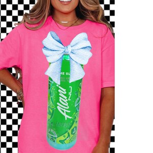 May include: A bright pink t-shirt featuring a green energy drink can with a light blue bow. The energy drink can has the brand name "Alani" in white script and the word "Energy" in green.