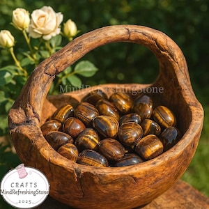 May include: A wooden basket filled with polished tiger's eye stones. The stones are a mix of brown and gold, with a striped pattern. The basket has a handle and is set against a blurred green background with white flowers. The text "MindRefreshingStone" is visible.