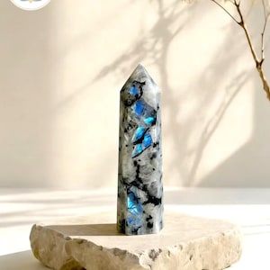 Puede incluir: Un obelisco de cristal pulido y alto con una base plana. El cristal es principalmente blanco y gris, con llamativas inclusiones azules. El obelisco se exhibe sobre una plataforma de piedra clara. El texto "MindRefreshingStone" es visible.