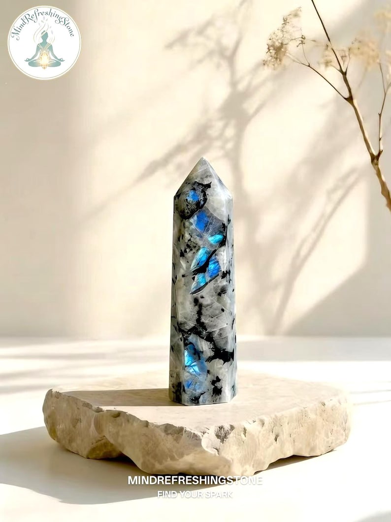 Puede incluir: Un obelisco de cristal pulido y alto con una base plana. El cristal es principalmente blanco y gris, con llamativas inclusiones azules. El obelisco se exhibe sobre una plataforma de piedra clara. El texto "MindRefreshingStone" es visible.