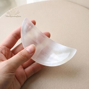 May include: A crescent moon-shaped object carved from translucent white selenite crystal with a smooth, polished surface. The stone features natural fibrous inclusions and a soft, pearlescent sheen. The text Mind Refreshing Stone is visible.