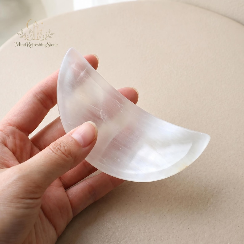 May include: A crescent moon-shaped object carved from translucent white selenite crystal with a smooth, polished surface. The stone features natural fibrous inclusions and a soft, pearlescent sheen. The text Mind Refreshing Stone is visible.