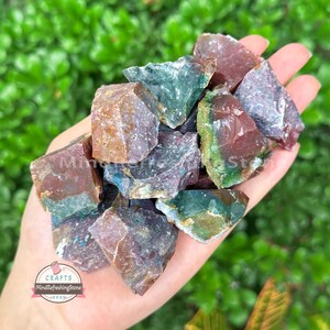 May include: A handful of raw, colorful gemstones in shades of red, green, and purple. The rough-cut stones are held in a hand, with a blurred green background. The image includes the text "MindRefreshingStone" and "Crafts 2025".