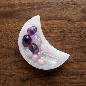 May include: A white crescent moon-shaped bowl containing a collection of polished gemstones, including deep purple amethyst, pale pink rose quartz, and a single clear, pointed quartz crystal point resting in the center.