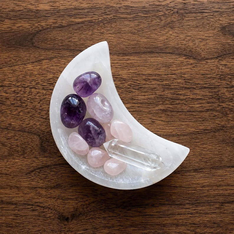 May include: A white crescent moon-shaped bowl containing a collection of polished gemstones, including deep purple amethyst, pale pink rose quartz, and a single clear, pointed quartz crystal point resting in the center.