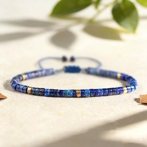 May include: A beaded bracelet featuring small, cylindrical lapis lazuli beads in various shades of blue, interspersed with metallic gold-toned spacer beads. The bracelet includes an adjustable cord closure with two dangling ends and a sliding knot.