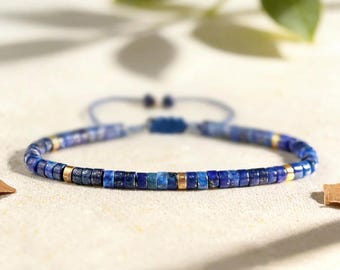 May include: A beaded bracelet featuring small, cylindrical lapis lazuli beads in various shades of blue, interspersed with metallic gold-toned spacer beads. The bracelet includes an adjustable cord closure with two dangling ends and a sliding knot.