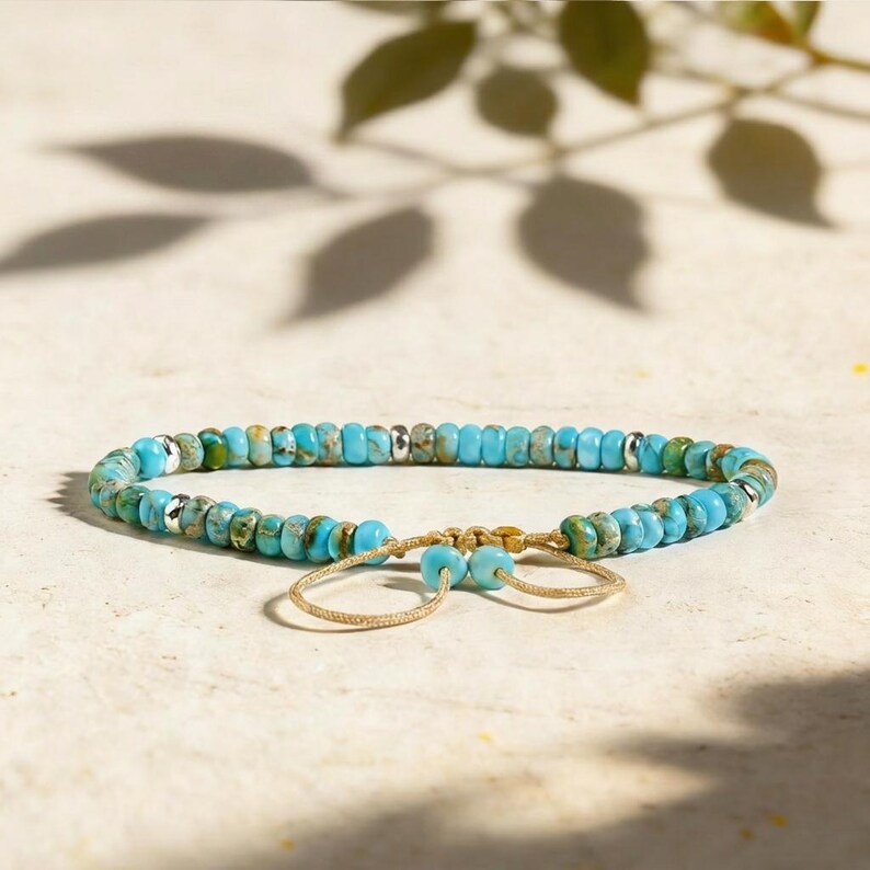 May include: A bracelet featuring round turquoise-colored stone beads with natural veining, interspersed with small metallic silver-toned spacer beads. The piece includes a tan cord closure with two dangling beads at the ends for adjustment.