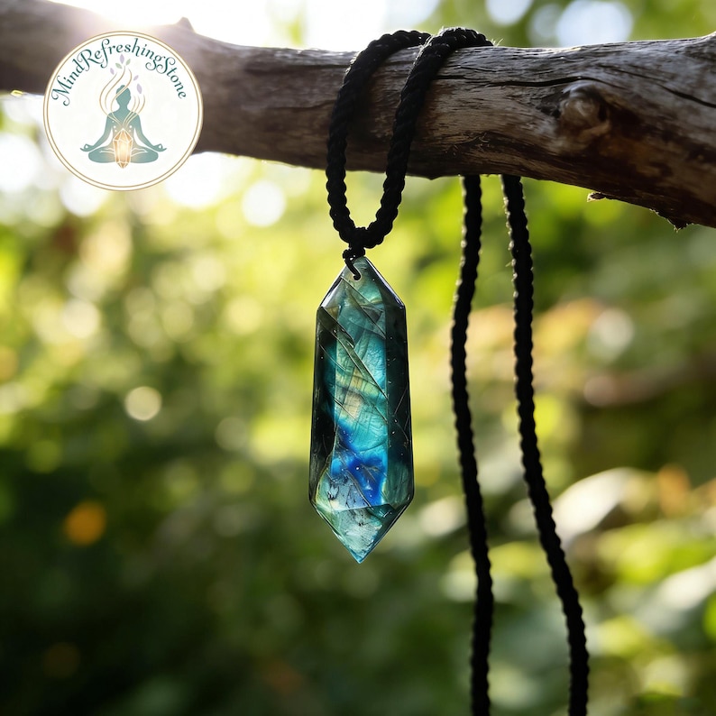 May include: A faceted, iridescent labradorite crystal pendant hangs from a black cord necklace. The crystal displays shades of blue, green, and yellow. The pendant is suspended from a black rope-like cord, and the background is blurred green foliage.
