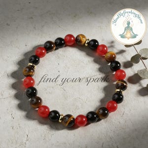 May include: A beaded bracelet featuring red, black, and tiger's eye beads, interspersed with gold-colored spacers. The bracelet is displayed on a neutral surface, with the words "find your spark" in cursive script. A circular logo is visible in the upper right corner.