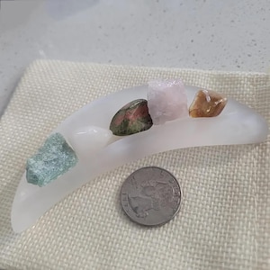 May include: A crescent-shaped white frosted glass dish holding four small raw gemstones, including green, pink, and brown varieties, positioned next to a silver United States quarter coin for scale on a textured beige fabric surface.