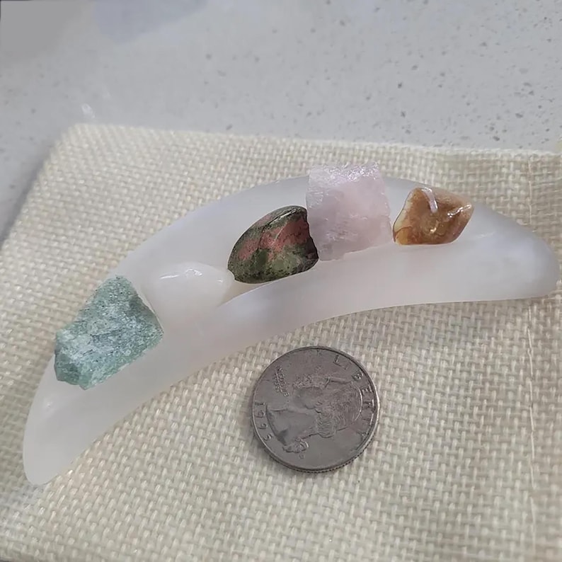 May include: A crescent-shaped white frosted glass dish holding four small raw gemstones, including green, pink, and brown varieties, positioned next to a silver United States quarter coin for scale on a textured beige fabric surface.