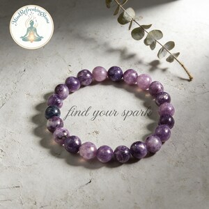 May include: A beaded bracelet featuring round, purple gemstones. The bracelet is displayed on a textured, light gray surface with the text "find your spark". A logo with a meditating figure is in the upper left corner.