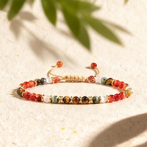May include: A beaded bracelet featuring alternating red, brown tiger eye, green, and pale pink beads separated by small gold-toned metal spacers. The bracelet includes an adjustable cord closure with two small red beads at the ends of the strings.