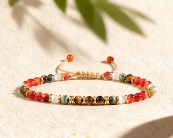 May include: A beaded bracelet featuring alternating red, brown tiger eye, green, and pale pink beads separated by small gold-toned metal spacers. The bracelet includes an adjustable cord closure with two small red beads at the ends of the strings.