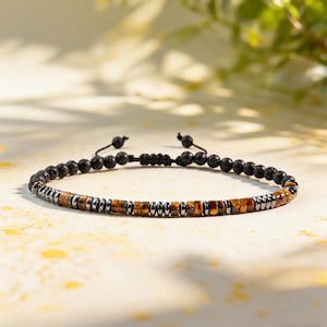 May include: A beaded bracelet featuring a mix of black porous lava stone beads, metallic silver-toned hematite discs, and golden-brown tiger eye stone beads. The bracelet includes an adjustable black cord closure with two small dangling beads.