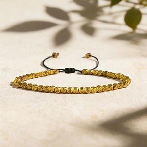 May include: A bracelet featuring a single strand of small, metallic gold-toned cube beads with etched circular patterns. The bracelet is secured with a black cord that includes an adjustable sliding knot closure and two small gold-toned end beads.