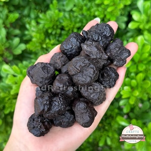 May include: A person's hand holds a collection of dark, textured stones, possibly hematite. The stones are a deep, matte black colour. The background is blurred green foliage, with the text "MindRefreshingStone" visible.