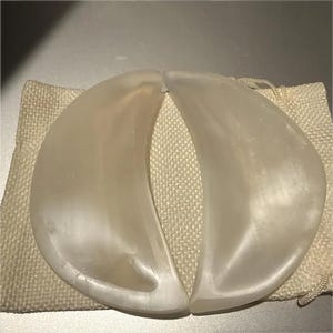 May include: Two crescent moon-shaped gua sha tools crafted from smooth, translucent white stone. The pieces feature curved edges and a polished finish, resting on a small, rectangular, off-white woven fabric drawstring pouch.