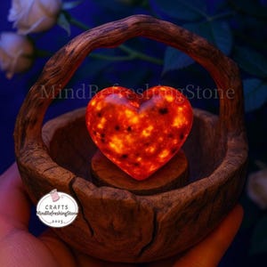 Puede incluir: Una piedra en forma de coraz&oacute;n, brillante, con un patr&oacute;n naranja y negro, colocada en una r&uacute;stica cesta de madera con asa. La cesta se sostiene en una mano, con un logo que dice "Crafts MindRefreshingStone 2025".