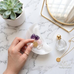 May include: A white crescent moon-shaped selenite bowl containing two purple amethyst stones, one pink rose quartz stone, one clear quartz stone, and a gold-toned circular pendant with an intricate filigree pattern.