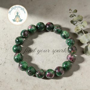 May include: A beaded bracelet featuring green and pink gemstone beads. The bracelet is displayed on a neutral surface with the words "find your spark" in script. A logo with a meditating figure is in the upper left corner.