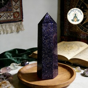 May include: A tall, six-sided obelisk made of dark blue goldstone with a shimmering, glitter-like appearance throughout. The stone features a pointed top and is positioned upright on a round, light brown wooden tray.