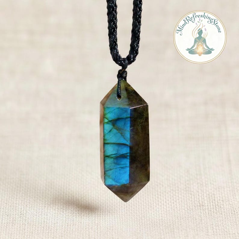 May include: A faceted labradorite crystal pendant with a black cord. The crystal has a mix of brown, blue, and green hues. The pendant is suspended from a braided black cord. A circular logo with the text "Mind Refreshing Stone" is in the upper right corner.