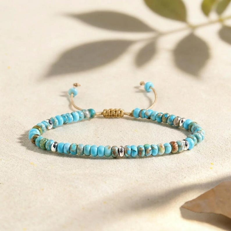 May include: A bracelet featuring a series of small, round, turquoise-colored beads with marbled brown and green patterns, interspersed with polished silver-toned metallic beads. The bracelet has a tan cord with an adjustable sliding knot closure.