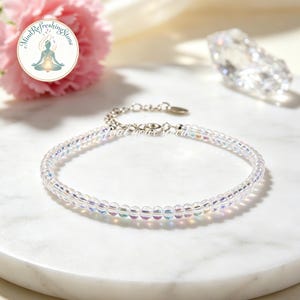 Op de afbeelding: Een delicate armband gemaakt van kleine, iriserende kralen. De armband heeft een zilverkleurige ketting en sluiting. De achtergrond bevat een roze bloem, een helder kristal en de tekst "Mind Refreshing Stone" en "Feed Your Spirit".