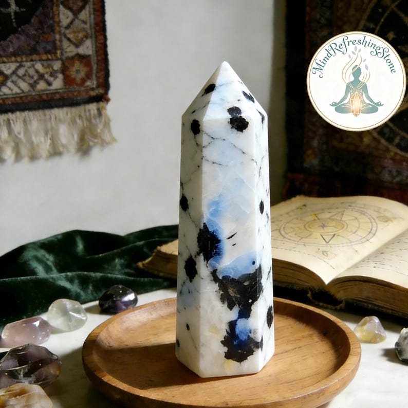 Puede incluir: Un obelisco de cristal alto, blanco y negro moteado, con detalles azules. La piedra pulida se exhibe en una bandeja de madera, con un libro y otros cristales en el fondo. El obelisco tiene una parte superior puntiaguda y una base hexagonal.