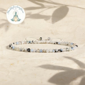 May include: A delicate bracelet featuring small, round, light blue, white, and black beads. The bracelet is strung on a white cord with adjustable sliding knots. The image includes a logo with the text "Mind Refreshing Stone".