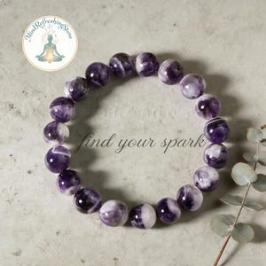 May include: A beaded bracelet made of round, polished amethyst stones. The stones are a mix of purple and white, strung together to form a bracelet. The text "find your spark" is written in cursive. A logo is in the upper left corner.