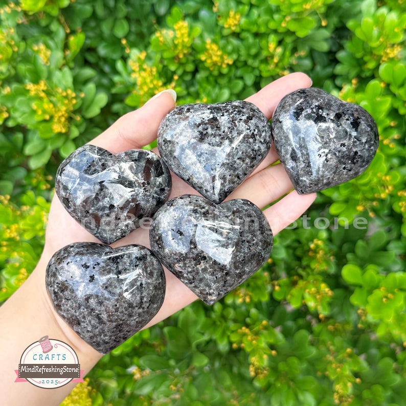 Puede incluir: Cinco piedras pulidas en forma de coraz&oacute;n con un patr&oacute;n moteado negro y gris. Las piedras se sostienen en una mano sobre un fondo de follaje verde. Las piedras tienen un acabado brillante y son suaves al tacto. La imagen incluye un logotipo que dice "Crafts MindRefreshingStone 2025".