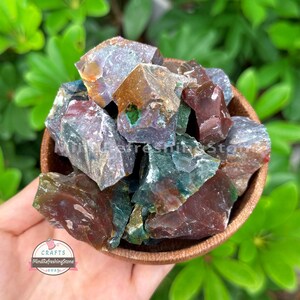 May include: A wooden bowl filled with raw, colorful gemstones. The stones display a mix of colors, including deep reds, greens, and purples. The bowl is held in a hand, with green foliage in the background. The text "CRAFTS MindRefreshingStone 2025" is visible.
