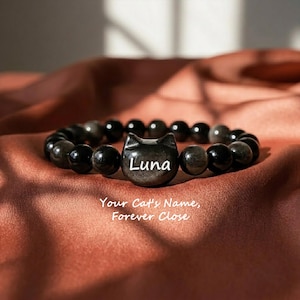 May include: A beaded bracelet featuring smooth, dark, reflective spherical beads and a central cat-shaped charm. The charm is engraved with the name "Luna" in white lettering. Below the bracelet, the text reads "Your Cat's Name, Forever Close."