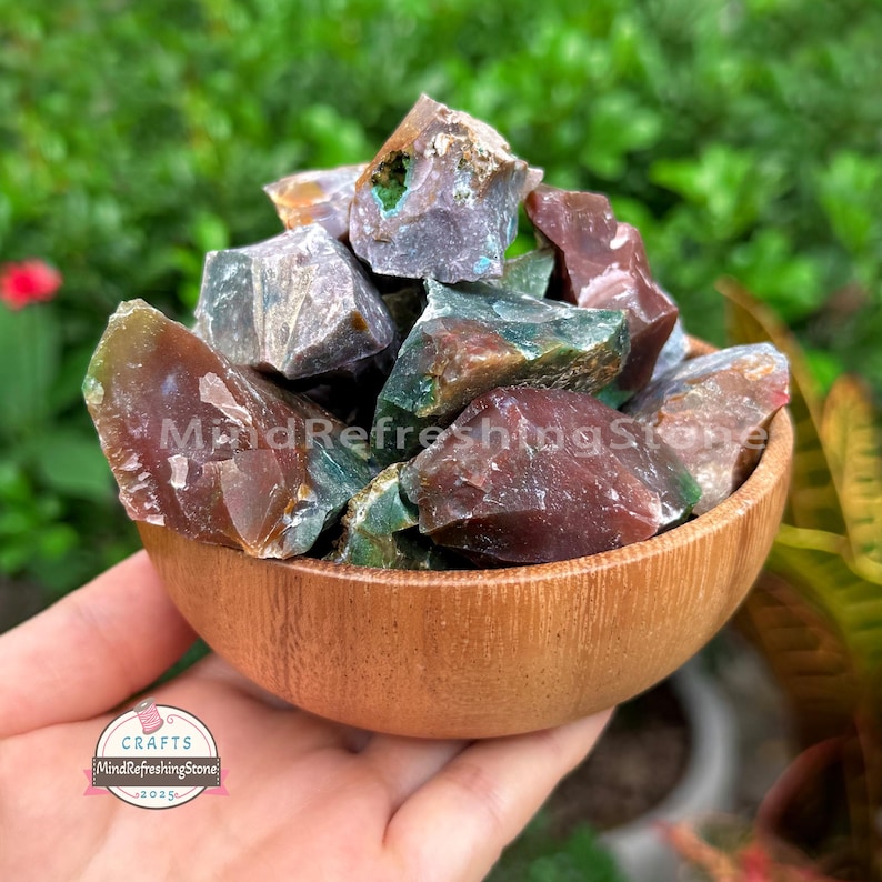 May include: A wooden bowl filled with raw, uncut gemstones in shades of green, brown, and gray. The bowl is held in a hand, with a blurred green background. The gemstones have a rough, natural texture. The bowl has a Crafts MindRefreshingStone 2025 sticker.