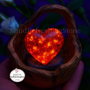 May include: A glowing red heart-shaped stone with yellow accents sits in a wooden basket. The basket has a handle and is made of a dark brown wood. The text "MindRefreshingStone" is visible.