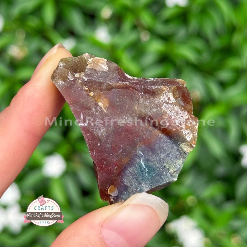 May include: A rough-cut gemstone with a mix of colors, including deep red, brown, and hints of blue and green. The stone is held in a hand, with the text "MindRefreshingStone" visible. The background is a blurred green.