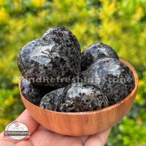 Puede incluir: Un cuenco de madera lleno de piedras pulidas en forma de coraz&oacute;n. Las piedras son una mezcla de gris oscuro y negro con un patr&oacute;n moteado. El cuenco se sostiene en una mano, con un fondo verde borroso.