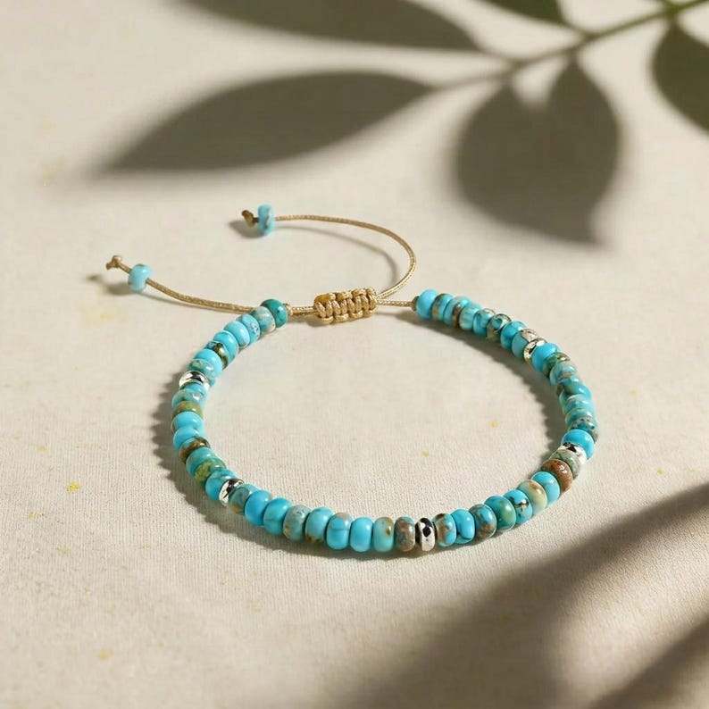 May include: A bracelet featuring small, round, turquoise-colored beads with mottled brown and green patterns, interspersed with polished silver-toned metal spacers. The piece includes a tan cord with an adjustable sliding knot closure.