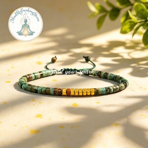 May include: A turquoise and gold beaded bracelet with the text "find your spark". The bracelet features small, round turquoise beads interspersed with gold and brown beads. The bracelet is on a light yellow surface.