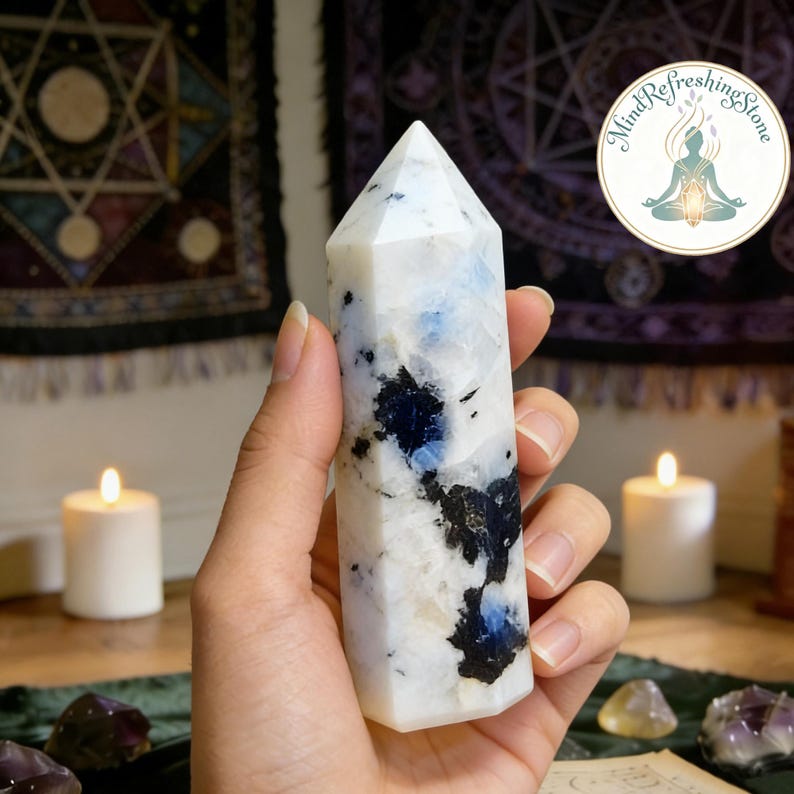 Puede incluir: Una torre de cristal de piedra lunar blanca y azul con inclusiones negras. La gema pulida se sostiene en una mano. El fondo incluye velas y un tapiz. La base es rectangular.