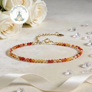May include: A delicate bracelet featuring small, round beads in shades of red, orange, and yellow, interspersed with gold-colored accents. The bracelet has an adjustable gold chain and is displayed on a white surface with decorative elements.