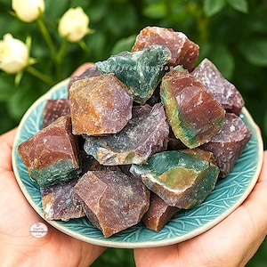 May include: A collection of raw, colorful gemstones in shades of red, green, and purple, displayed in a teal-colored ceramic bowl. The gemstones have a rough, natural texture and are held in a person's hands. The bowl has a decorative, embossed pattern.