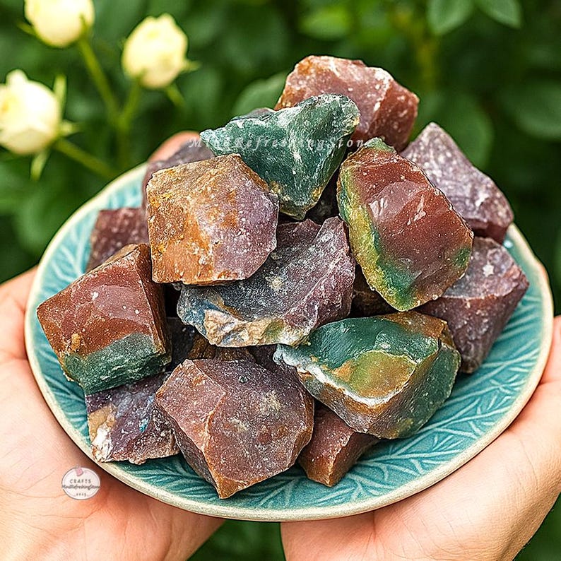 May include: A collection of raw, colorful gemstones in shades of red, green, and purple, displayed in a teal-colored ceramic bowl. The gemstones have a rough, natural texture and are held in a person's hands. The bowl has a decorative, embossed pattern.