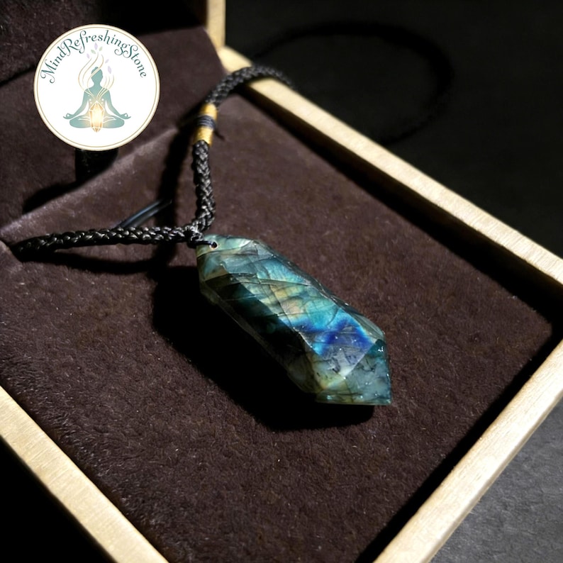 May include: A faceted labradorite pendant with a black cord necklace. The gemstone displays iridescent blue, green, and yellow hues. The pendant is displayed in a brown box. The image includes a circular logo with the text "Mind Refreshing Stone".