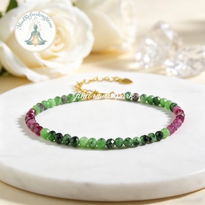 Pode incluir: Uma delicada pulseira de contas com pedras preciosas verdes e granadas alternadas. A corrente dourada é ajustável. A pulseira é exibida em uma superfície branca com o texto "find your spark".